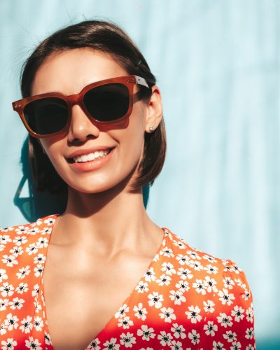 Young beautiful smiling female in trendy summer red dress. Sexy carefree woman posing near blue wall in studio. Positive model having fun. Cheerful and happy. At sunny day. Shadow from window